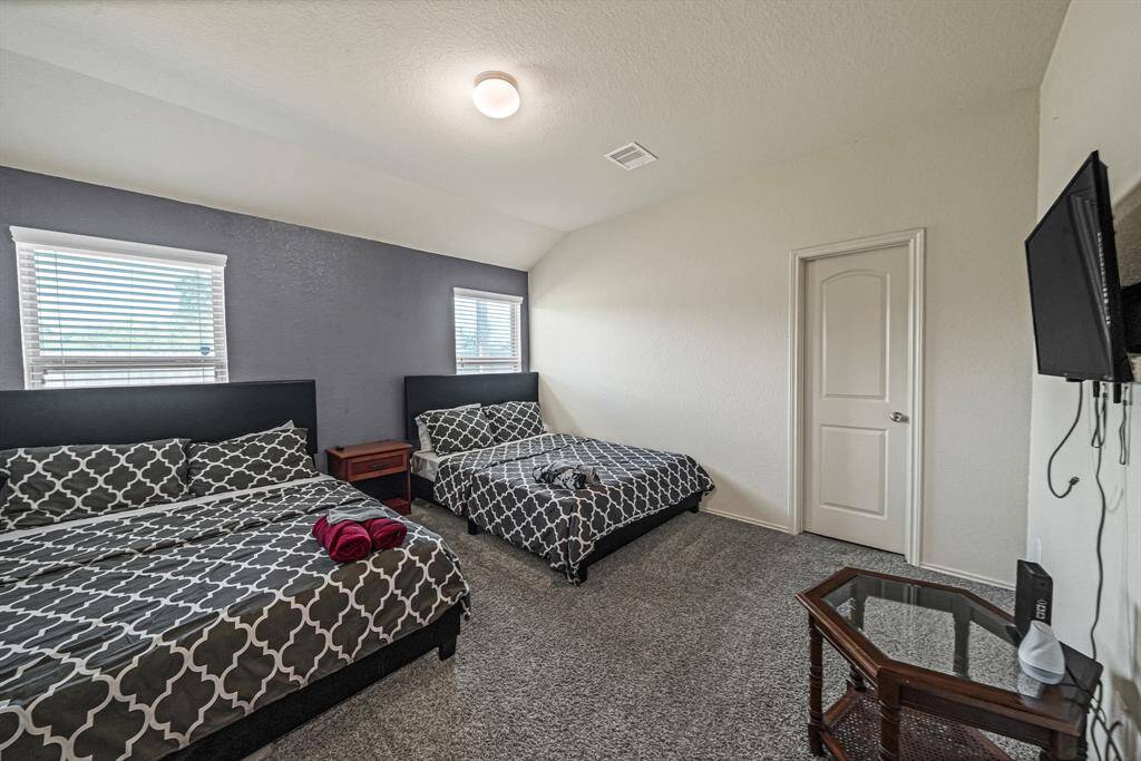 This spacious bedroom features two large beds with patterned bedding, a modern light fixture, and neutral walls. It includes a mounted TV, two small windows for natural light, and a carpeted floor for comfort.