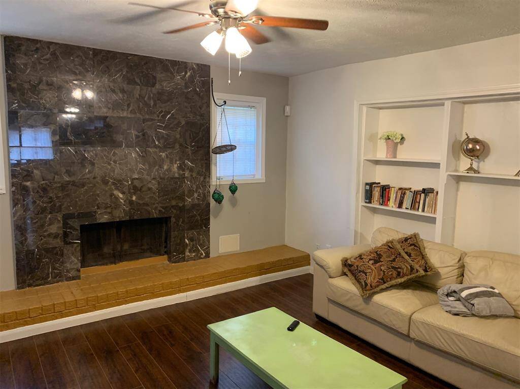 This cozy living room features a striking dark stone fireplace, a ceiling fan, and built-in shelves for storage. The space includes a comfortable beige sofa and hardwood flooring, offering a warm and inviting atmosphere.