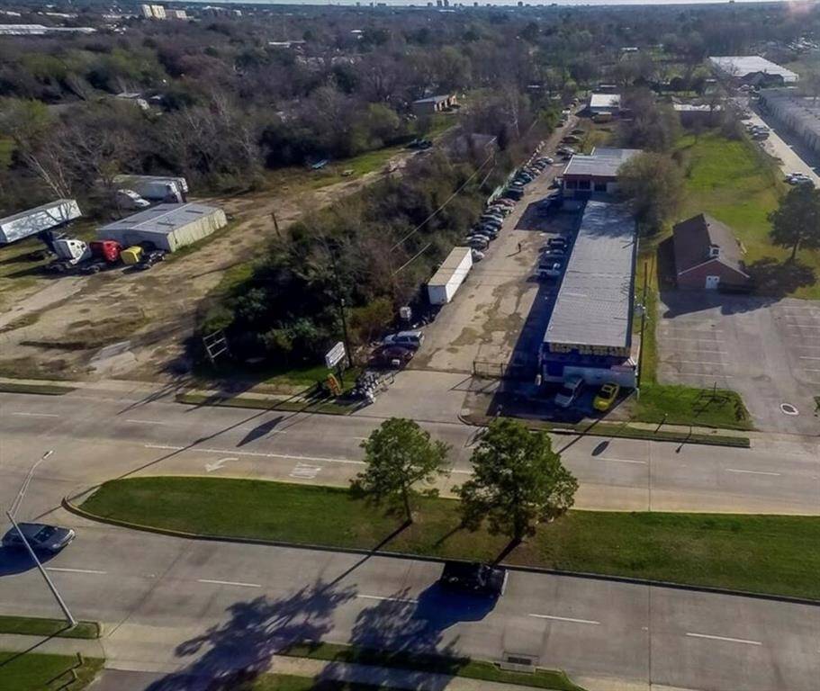 This aerial view features a long commercial building with ample parking, set in a spacious area alongside a wide road. Surrounded by open land and additional structures, it offers excellent development potential amid a blend of greenery and infrastructure.