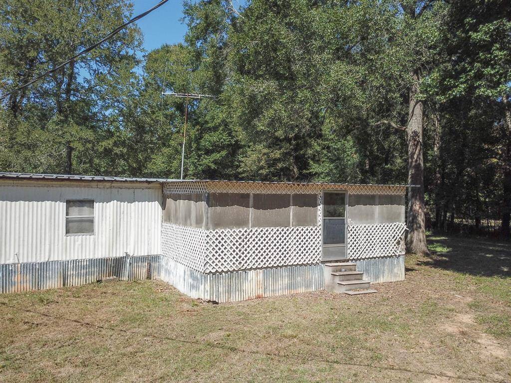 A small, modest house with a corrugated metal exterior and white lattice skirting, surrounded by trees under a clear sky.