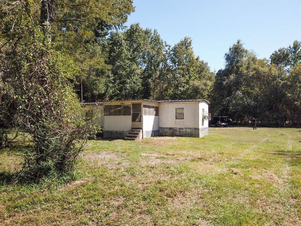 An old, weathered mobile home sits in a grassy field with trees around, under a clear sky. A person and vehicle are visible in the background.