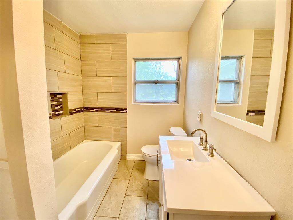 Remodeled modern bathroom featuring a white bathtub with beige tile surround, a single sink vanity, and a toilet. Natural light comes from a window above the tub. The space has a clean and bright aesthetic.