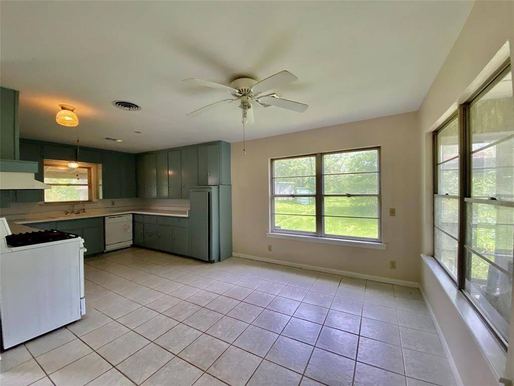 The kitchen with ample natural light, featuring tile flooring, green cabinetry, and a ceiling fan. It includes a window over the sink and a large window on the side wall, providing a view of the greenery outside.