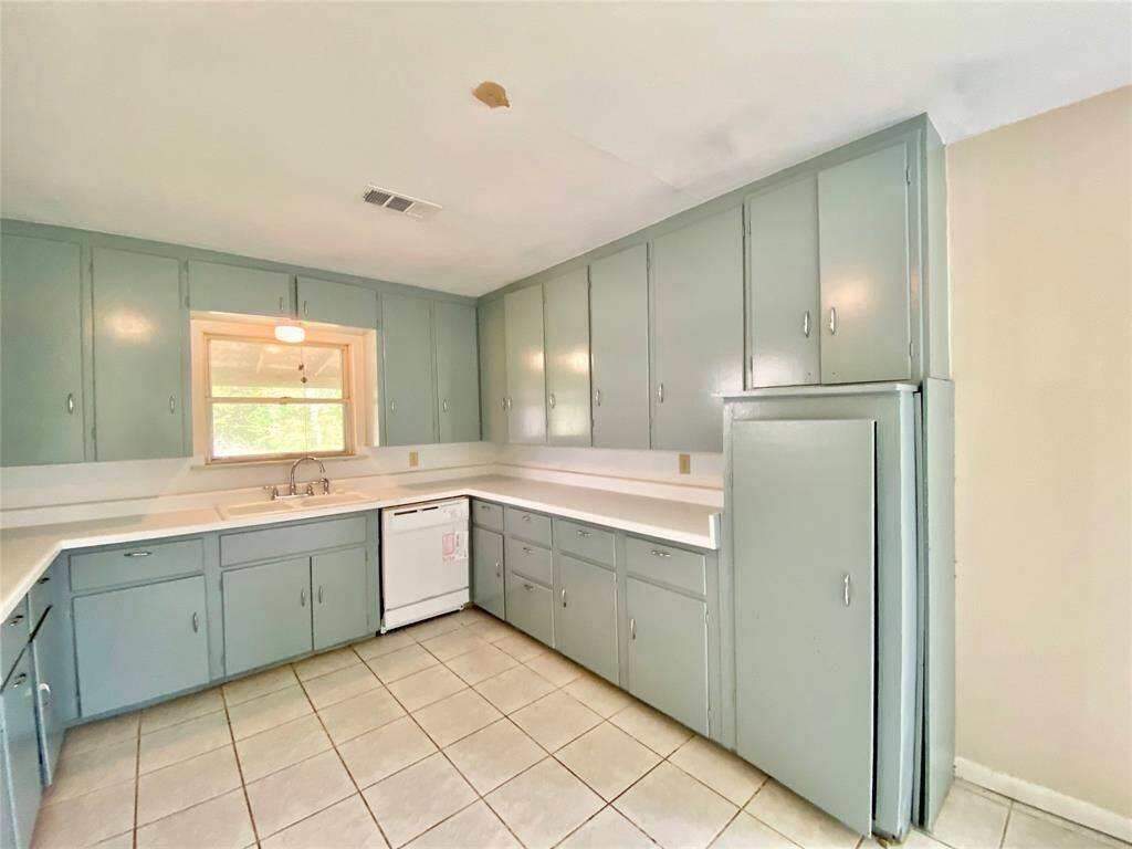 This kitchen features light blue cabinetry with ample storage, tile flooring, a window above the sink providing natural light, and includes a white dishwasher and a full-sized refrigerator.