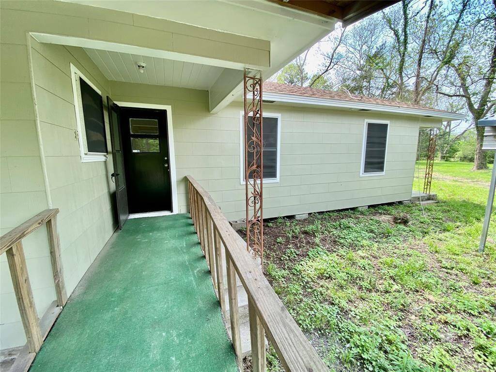 One-story home featuring a covered entryway, green carpeted porch with wooden railings, and a view of the yard with mature trees.