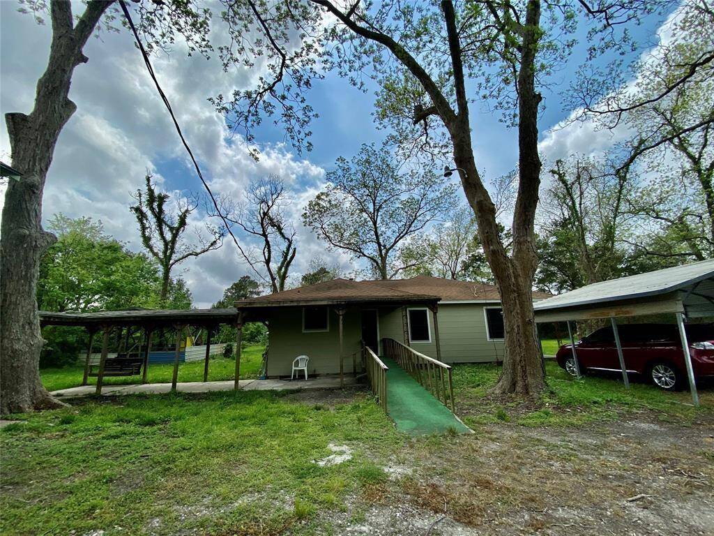 This is a single-story home with a green roof and beige siding, featuring a carport on the right and a covered porch area on the left. The property is surrounded by mature trees and has a natural, grassy yard.