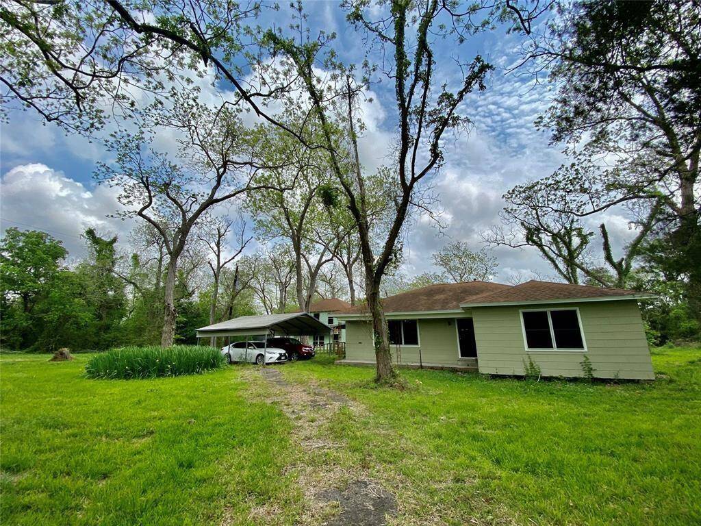 One-story home with a green exterior and white trim, featuring a carport on the left side. The property is surrounded by mature trees and has ample green yard space.
