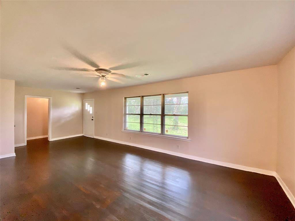 A bright, spacious living room with hardwood floors,  neutral paint, and a large window allowing for ample natural light. It features a ceiling fan and offers a clean, blank canvas for personalization.