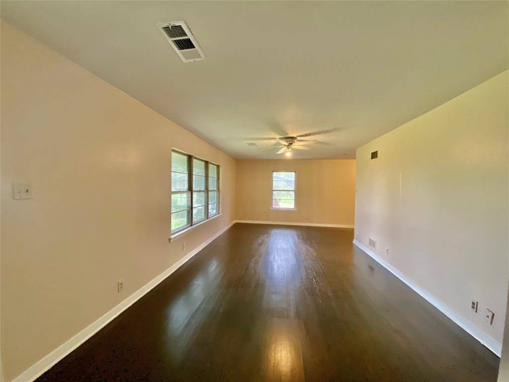 Spacious, well-lit room with gleaming hardwood floors, neutral wall colors, and a ceiling fan. There are multiple windows allowing for plenty of natural light.
