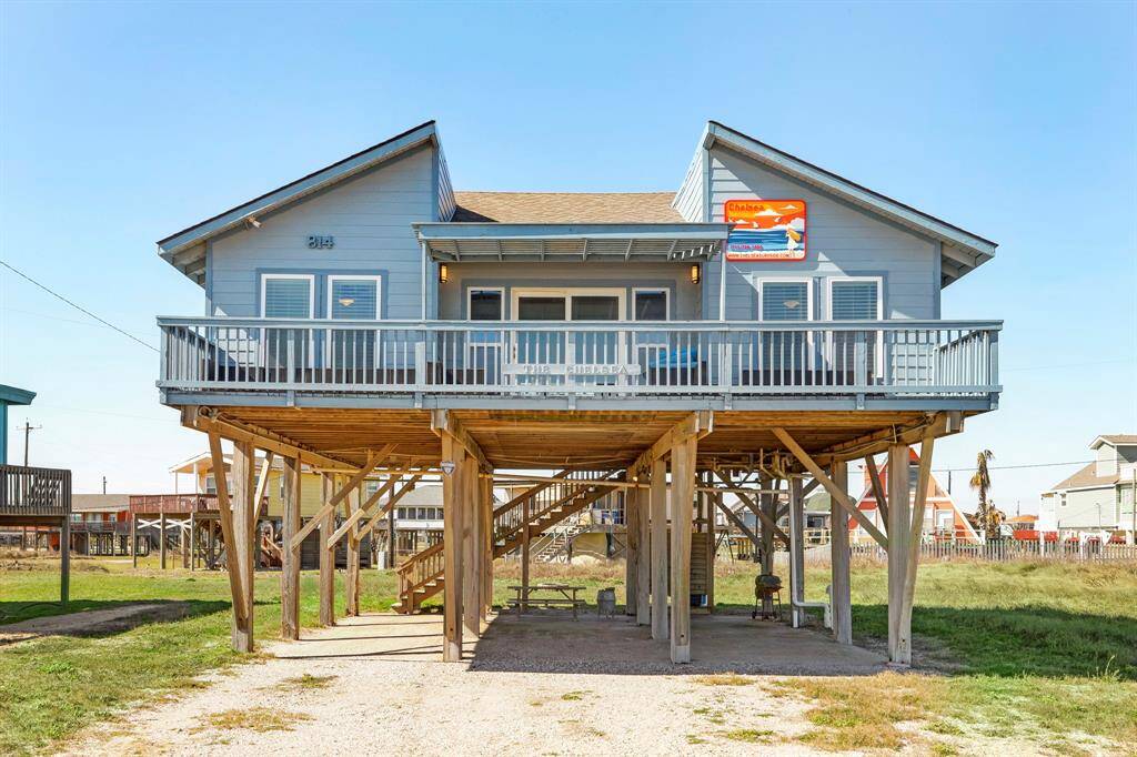 Elevated beach house with a spacious front deck, offering ample outdoor space and views. Features include a light blue exterior, large windows, and a covered area underneath for parking or storage. Ideal for coastal living.