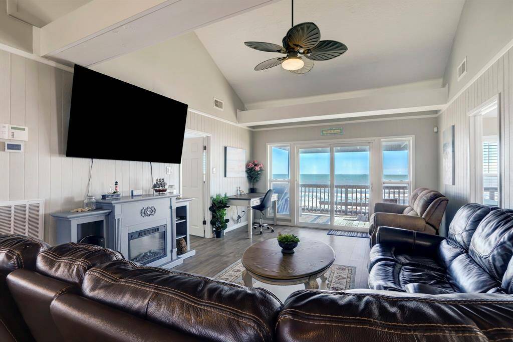 This inviting living room features a vaulted ceiling, leather seating, a large wall-mounted TV, and a cozy fireplace. The space offers stunning ocean views through expansive glass doors leading to a balcony.