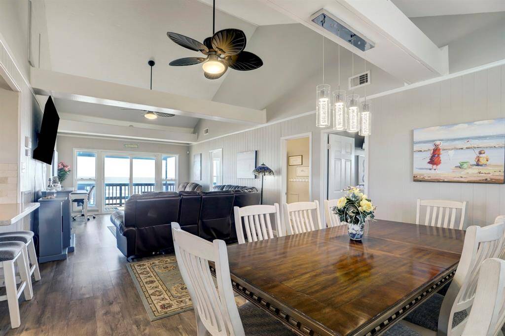 This spacious, open-concept living and dining area features high ceilings, modern lighting, and a large window with ocean views. The room is elegantly decorated with a sleek ceiling fan, plush seating, and a large dining table, creating an inviting atmosphere for relaxation and entertaining.