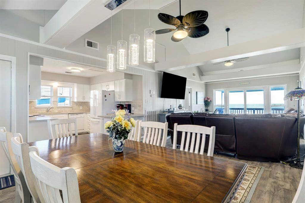 This open-concept living space features a modern kitchen with white cabinetry and a cozy dining area. The living room includes a large sectional, ceiling fans, and a wall-mounted TV, leading to a balcony with ocean views, perfect for relaxation and entertaining.
