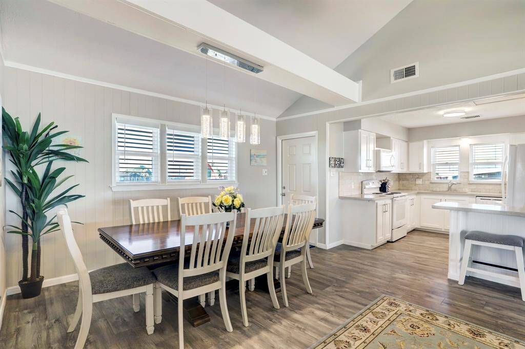 This photo showcases a bright, open-concept dining and kitchen area with high ceilings. The space features modern lighting, a large dining table, and a breakfast bar. The kitchen is equipped with white cabinetry, a tiled backsplash, and ample natural light from multiple windows.