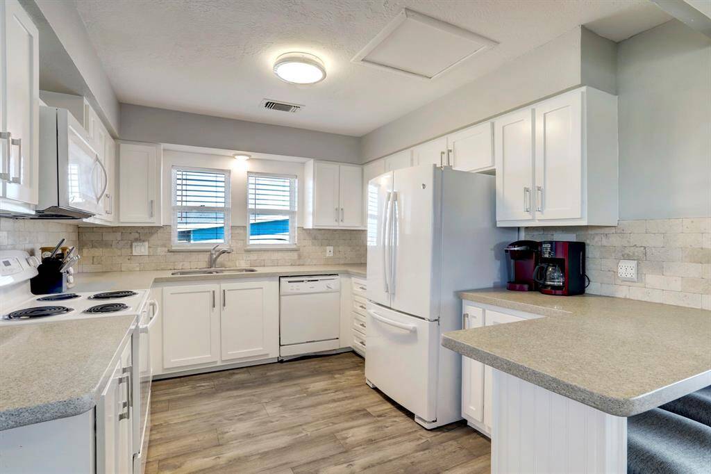 Bright, modern kitchen with white cabinetry, stone backsplash, and light countertops. Includes appliances like a fridge, stove, and dishwasher. Features wood-style flooring and ample natural light from two windows.