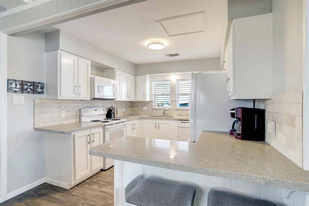 Bright and modern kitchen with white cabinetry, light countertops, and a breakfast bar. Features include stainless steel appliances, a tiled backsplash, and ample natural light from the windows.