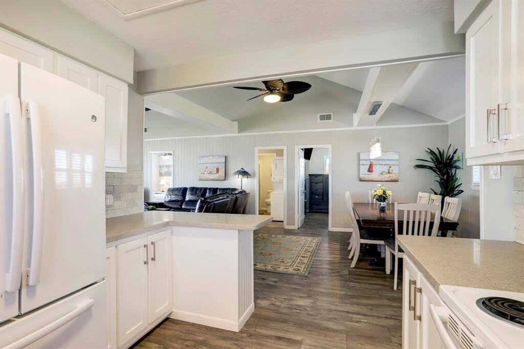 This photo showcases a bright, open-concept kitchen and living area with vaulted ceilings and modern furnishings. The kitchen features white cabinets and countertops, while the adjacent dining area has a cozy, inviting atmosphere with a ceiling fan and decorative touches.