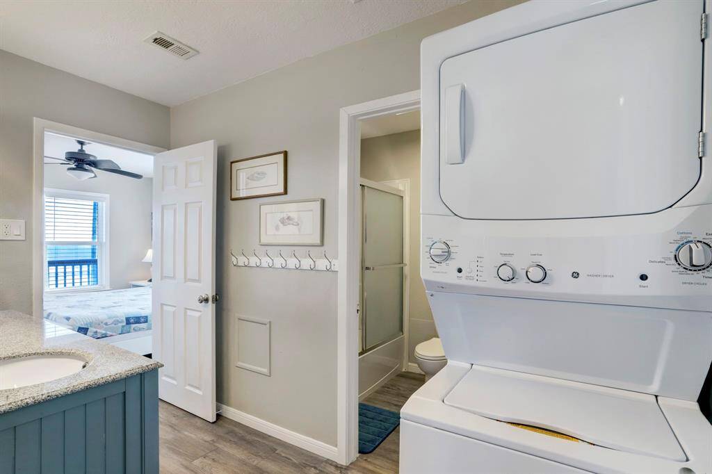 This photo shows a compact laundry area with a stackable washer and dryer unit, adjacent to a bathroom with a shower. The space features modern decor, hooks for hanging items, and leads into a bedroom with natural light.