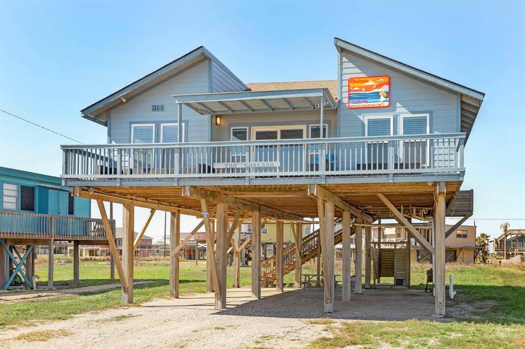 This is a charming elevated beach house with a spacious wrap-around deck, perfect for enjoying coastal views. The house is supported by sturdy wooden stilts, offering covered parking underneath. Its light blue exterior and modern design create a welcoming seaside retreat.