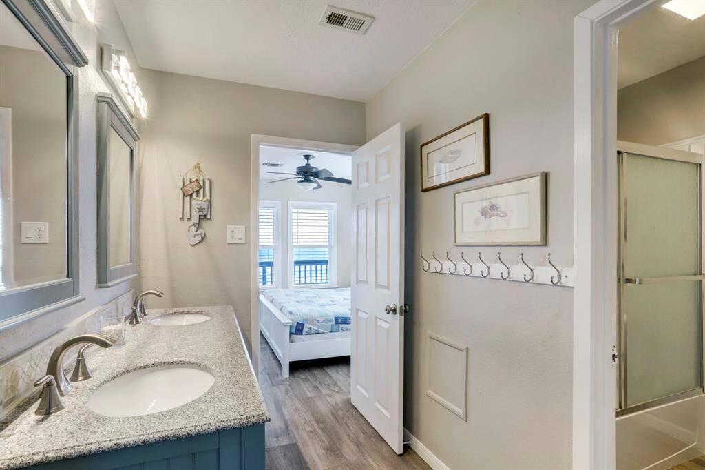 Bright bathroom with dual sinks, granite countertops, and modern fixtures. Leads into a bedroom with wood flooring and natural light through large windows.