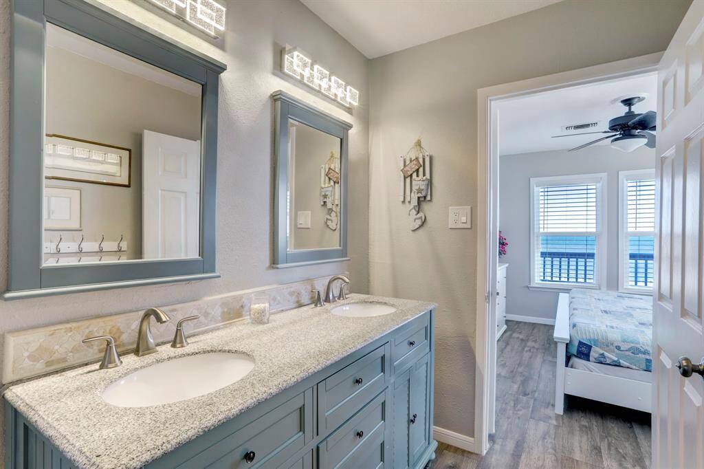 This photo shows a bright bathroom with a double vanity featuring granite countertops and stylish mirrors. The room leads into a bedroom with large windows and a ceiling fan, offering a cozy and modern feel.