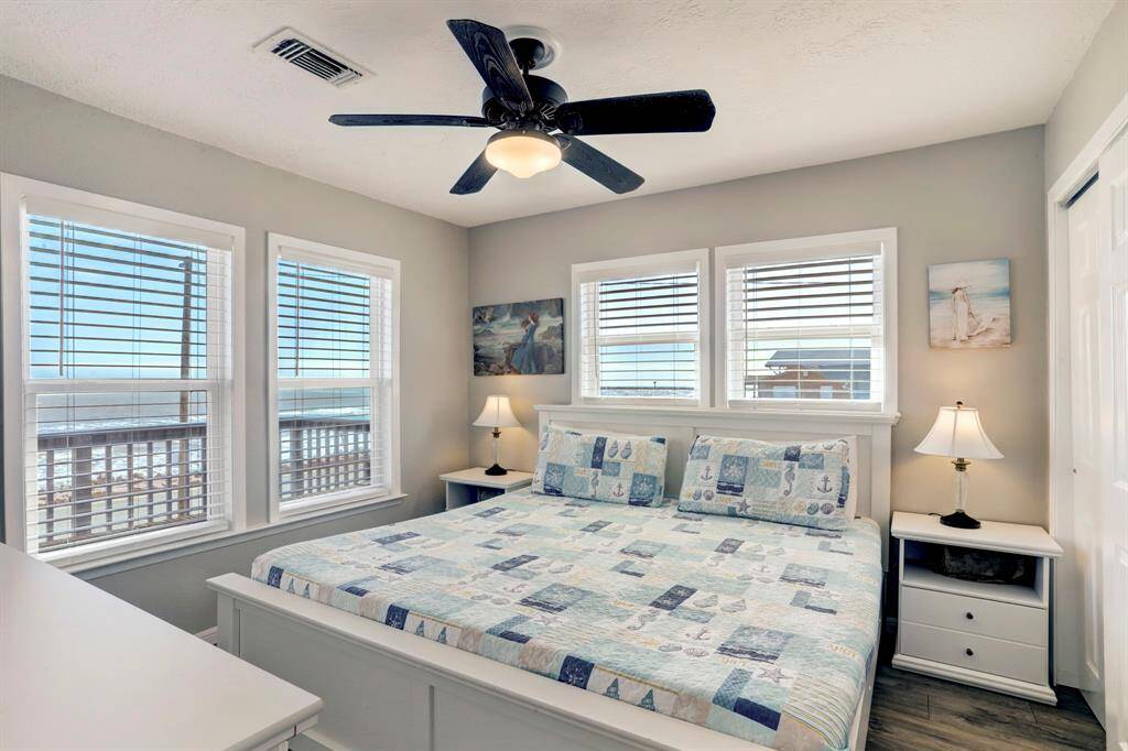 Bright bedroom with ocean views, featuring large windows, a ceiling fan, and coastal-themed decor. The room includes a spacious bed with a quilted cover, two bedside tables with lamps, and a relaxing, airy atmosphere.
