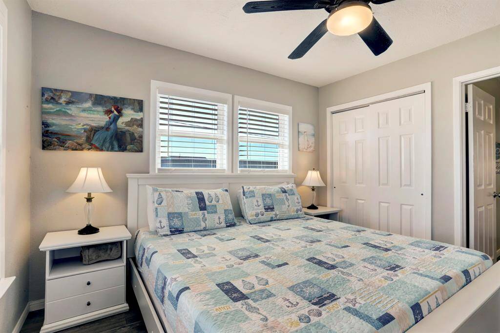 A cozy bedroom with a nautical-themed quilt on a large bed, dual bedside lamps, and a ceiling fan. It features a closet with sliding doors and artwork on the walls, creating a serene atmosphere.