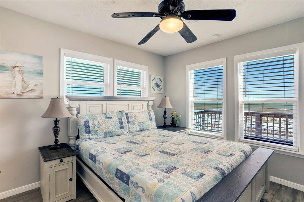 This bright bedroom features a cozy bed with patterned bedding, flanked by two stylish nightstands and lamps. Large windows offer stunning ocean views, while a ceiling fan and light-colored walls enhance the airy feel.