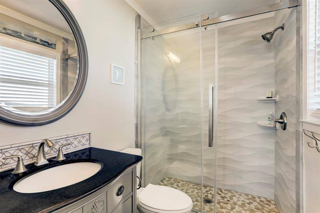 Modern bathroom featuring a sleek glass-enclosed shower with textured tiles. It has a stylish vanity with a black countertop and decorative backsplash, complemented by a round mirror and elegant fixtures. Natural light streams in through a window with blinds.
