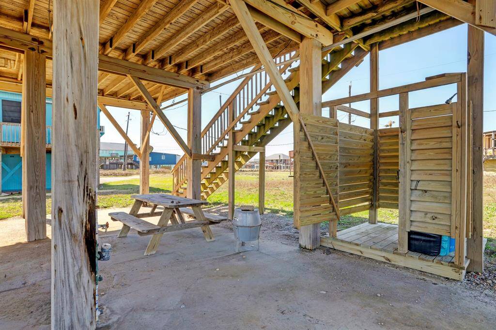 This image shows an outdoor space under a raised beach house featuring a wooden staircase, picnic table, and a small enclosed outdoor shower area. It's ideal for casual gatherings and rinsing off after a day at the beach.