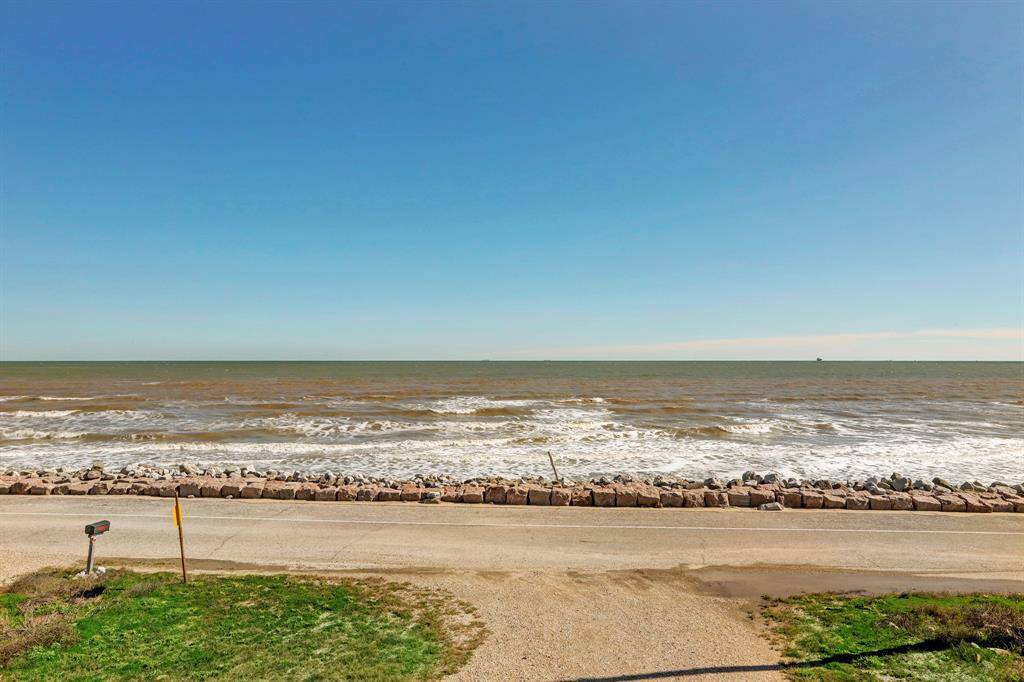This photo showcases a serene ocean view from a coastal property, featuring a clear blue sky, gentle waves, and a rocky shoreline adjacent to a quiet road, ideal for beachside living.