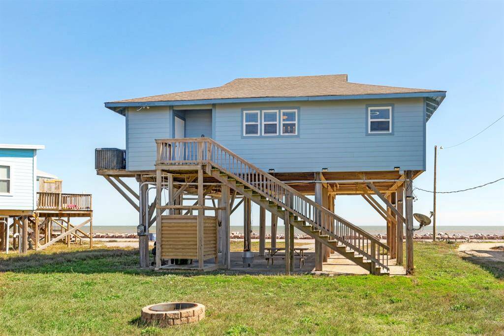 This elevated coastal home features a charming light blue exterior with a spacious wooden deck and stairs. It's positioned near the shoreline, offering scenic ocean views and a grassy yard, ideal for beachside living.