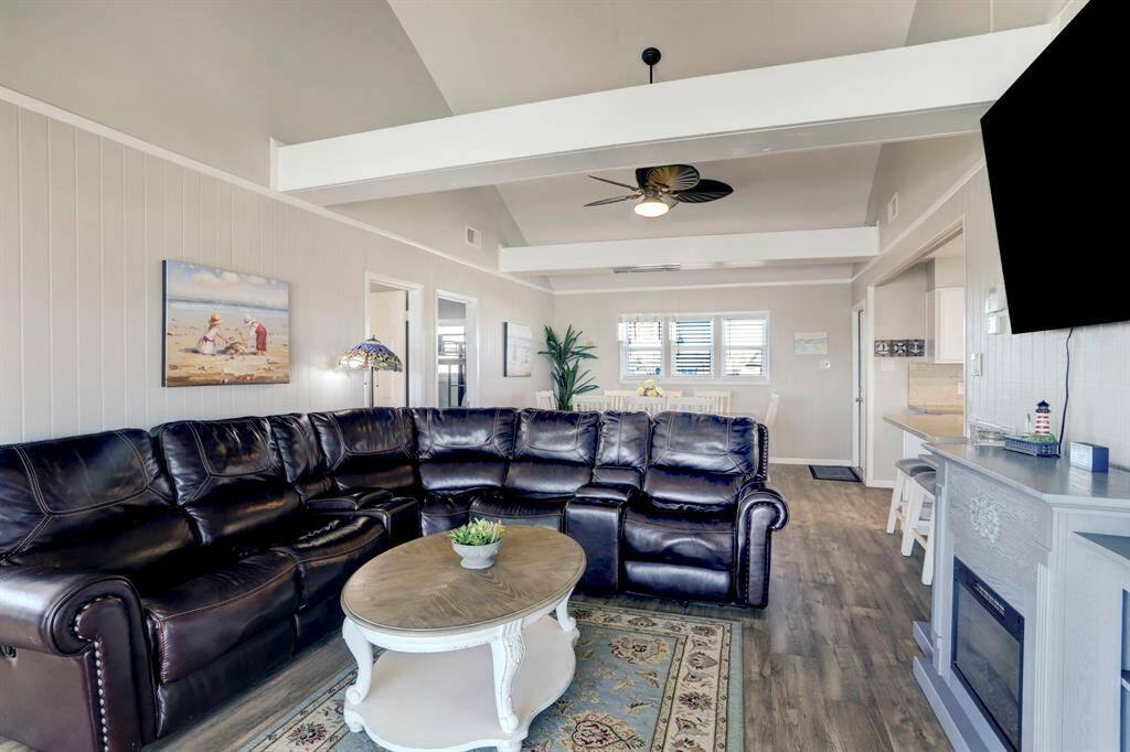Bright and inviting living area featuring a large L-shaped leather sofa, modern ceiling fan, and a wall-mounted TV. The open layout flows into a cozy dining space with natural light, hardwood floors, and neutral tones, creating a comfortable and stylish setting.