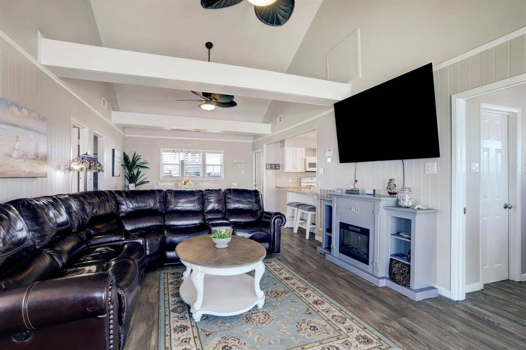 Spacious living room with vaulted ceilings, featuring a large sectional sofa, wall-mounted TV, and electric fireplace. The open layout flows into a bright kitchen with a breakfast bar, creating a cozy and inviting atmosphere.