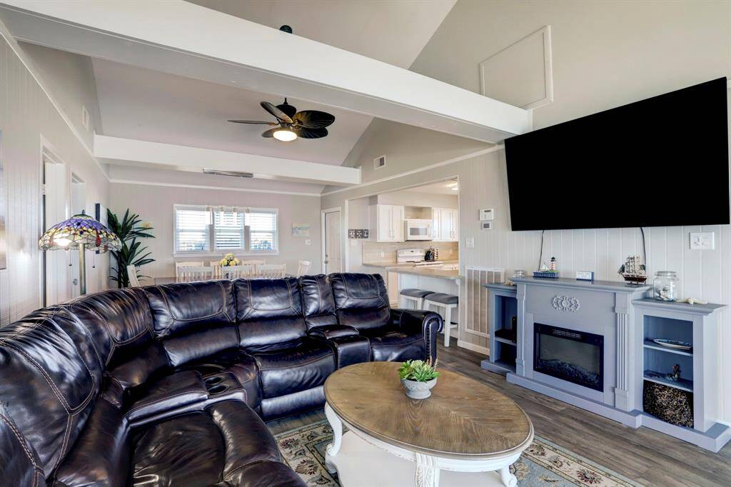 This inviting living room features a vaulted ceiling, large sectional sofa, and a wall-mounted TV above a decorative fireplace. The open layout leads to a bright dining area and a modern kitchen with bar seating, creating a spacious and welcoming environment.