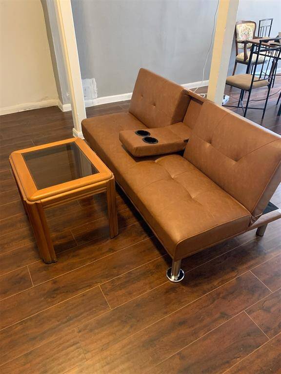 This is a modern living area featuring a tan sofa with sleek metal legs and a built-in cup holder, paired with a wooden side table with glass top. The room has dark hardwood floors and a neutral wall color, which could complement a variety of decor styles.