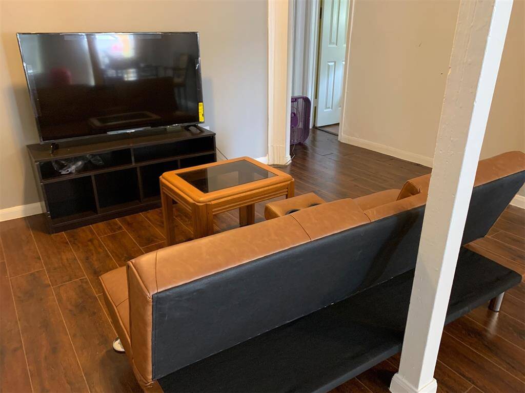 This is a cozy living room space featuring a dark wooden floor, a modern flat-screen TV on a black stand, a brown leather sofa, and a wooden coffee table with a glass top. The room is well-lit and leads to another room through an open doorway, with a white structural column adding character to the space.