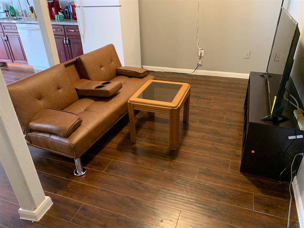 This room features a modern L-shaped leather sofa and a simple wooden coffee table on a dark hardwood floor. The space is open to the kitchen, with a view of wooden cabinets and appliances. A flat-screen TV is positioned at the edge, and the walls are painted in a neutral color.