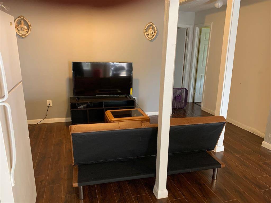 This is a living space featuring hardwood floors, with a view into another room. There's a black sofa and a flat-screen TV on a stand. The room is lit with natural light and has decorative wall sconces. A structural column partially obstructs the space.