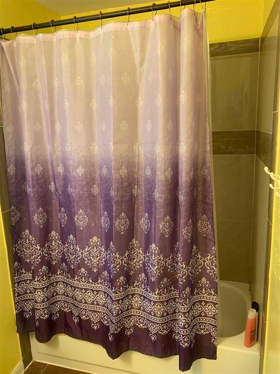 The photo shows a bathroom with a purple shower curtain featuring decorative patterns, set against a yellow wall and a tiled shower area.