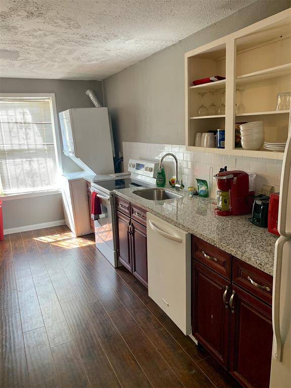 This is a cozy kitchen featuring wood-look flooring, cherry wood cabinets, white appliances, and a clean white tile backsplash. Natural light streams in from the window over the sink.