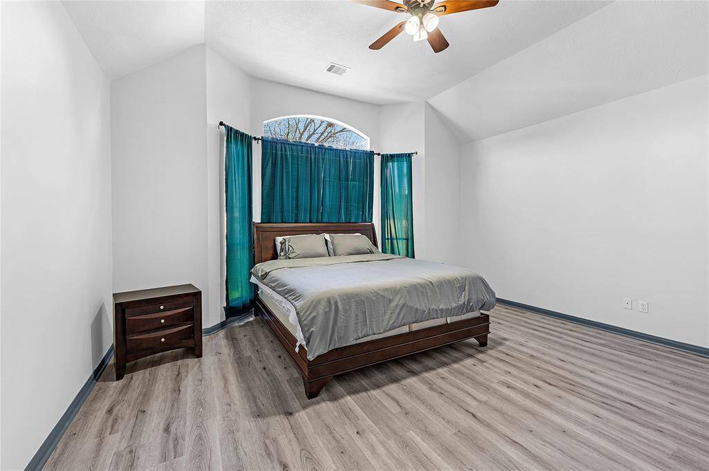 This bedroom features a vaulted ceiling with a ceiling fan, a large window with teal curtains, and wood-look flooring. It includes a double bed with a wooden headboard and a matching nightstand, creating a cozy and spacious atmosphere.