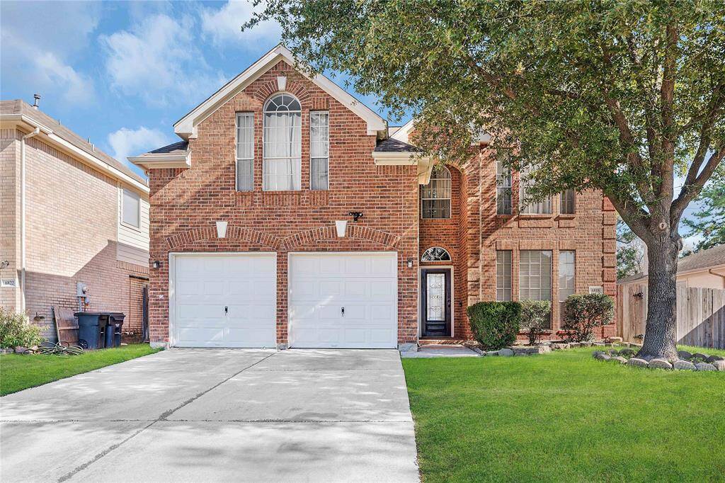This is a two-story brick home with a double garage and a well-maintained front yard. It features large windows, a prominent entryway, and a mature tree providing shade. The driveway is spacious and the overall curb appeal is inviting.