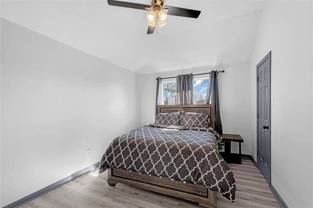 This cozy bedroom features a comfortable bed with a patterned duvet, modern ceiling fan, and natural light from a window with gray curtains. The light wood flooring adds warmth and complements the neutral wall tones.