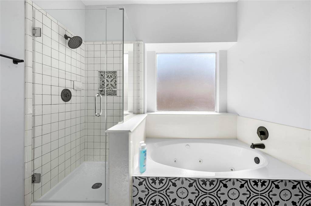This bathroom features a spacious glass-enclosed shower with classic white tiling and a stylish accent tile. Adjacent is a luxurious corner jetted bathtub with decorative black and white tile detailing, complemented by a frosted window for natural light and privacy.