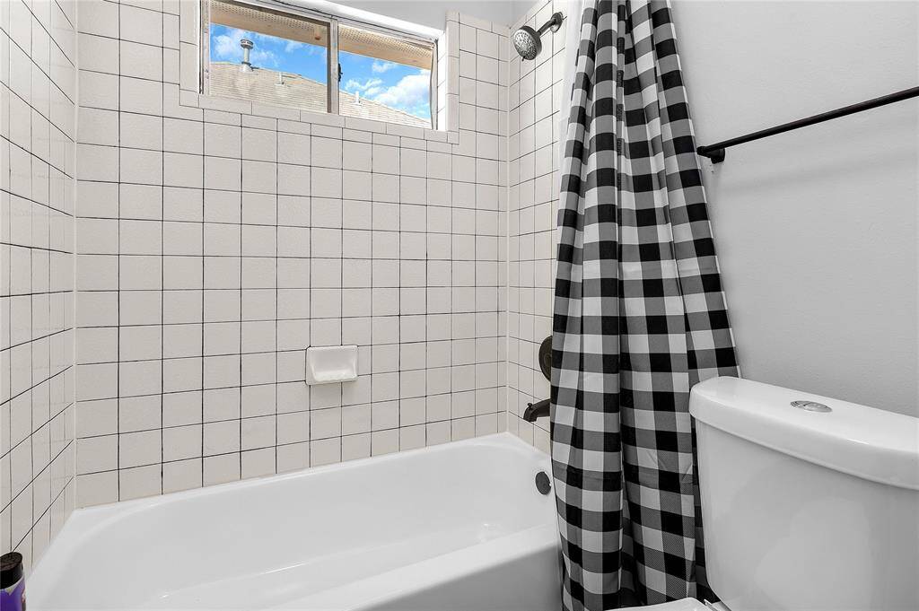 Compact bathroom featuring a tiled shower with a checkered curtain, a white bathtub, and a toilet. A window above the shower allows natural light in.