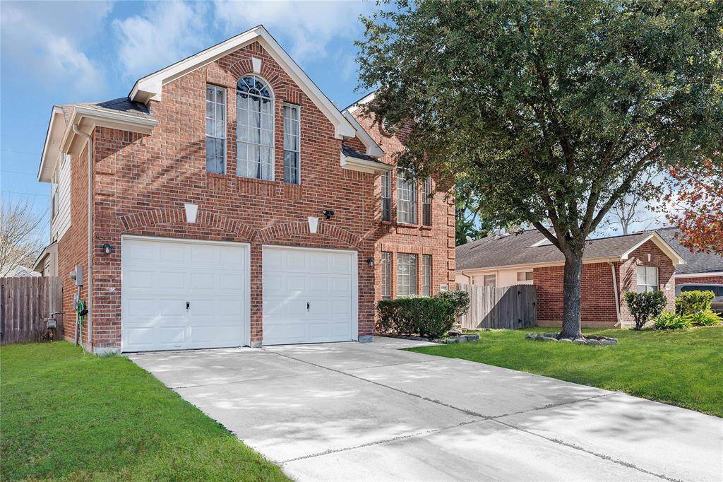 This photo features a charming two-story brick home with a double garage. The front yard is well-maintained with a large tree and lush greenery. The driveway is spacious, and the home has an elegant arched window above the garage, adding to its curb appeal.