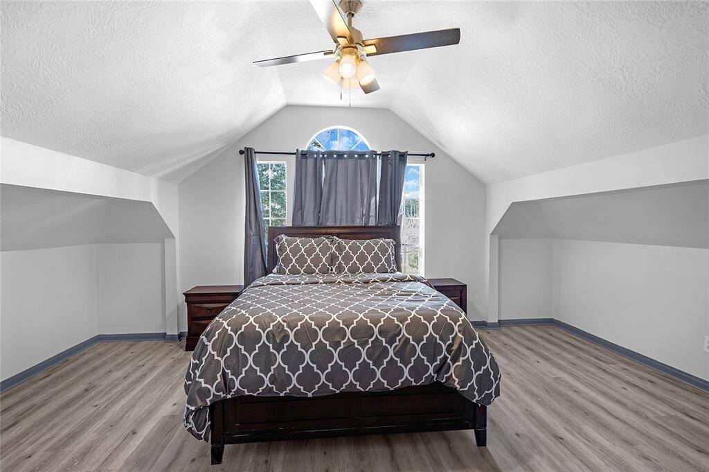 This cozy bedroom features a vaulted ceiling, a central light fixture with a fan, and a large arched window for natural light. It has a modern wood-look floor and neutral walls, creating a spacious and inviting atmosphere.