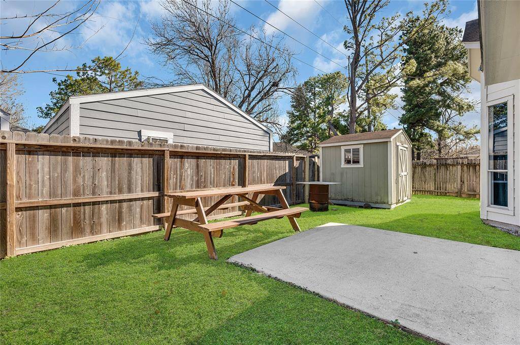 This backyard features a well-maintained lawn with a wooden picnic table, a small storage shed, and a concrete patio area. It's enclosed by a wooden fence, providing privacy and a cozy outdoor space.