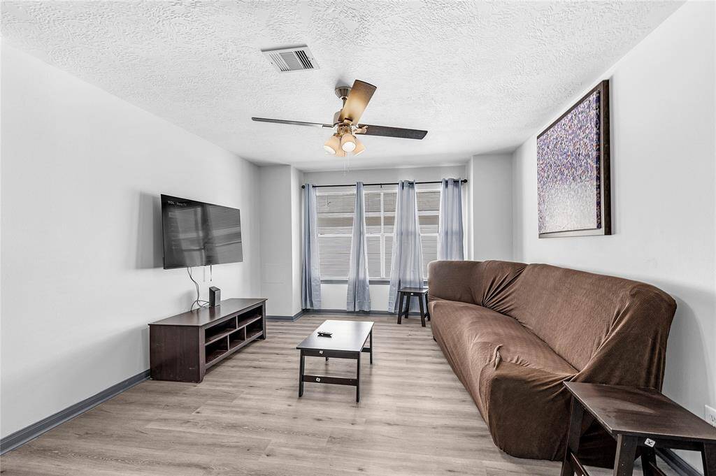This living room features a modern design with a ceiling fan, large wall-mounted TV, and a comfortable brown sofa. It has wood-style flooring and large windows with sheer curtains, creating a bright and inviting space.